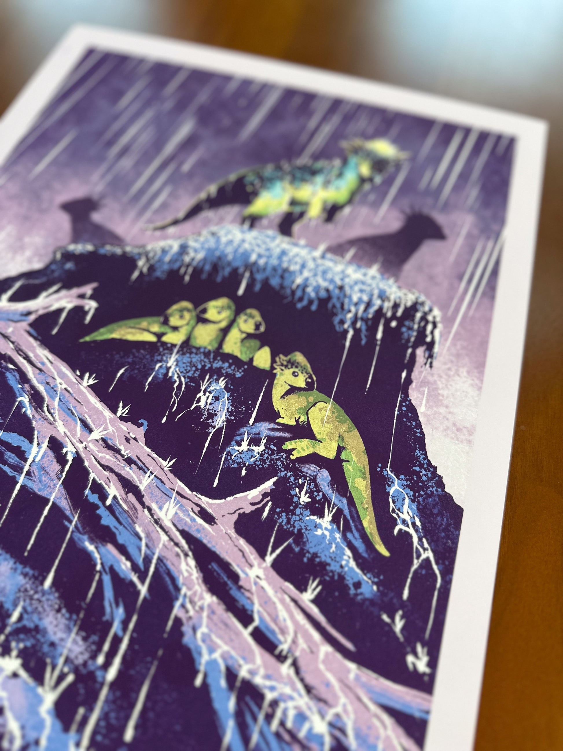 A close-up of an art print featuring a stylized illustration of small dinosaurs on a rainy cliffside under a purple rainy sky, photographed on a wooden surface.