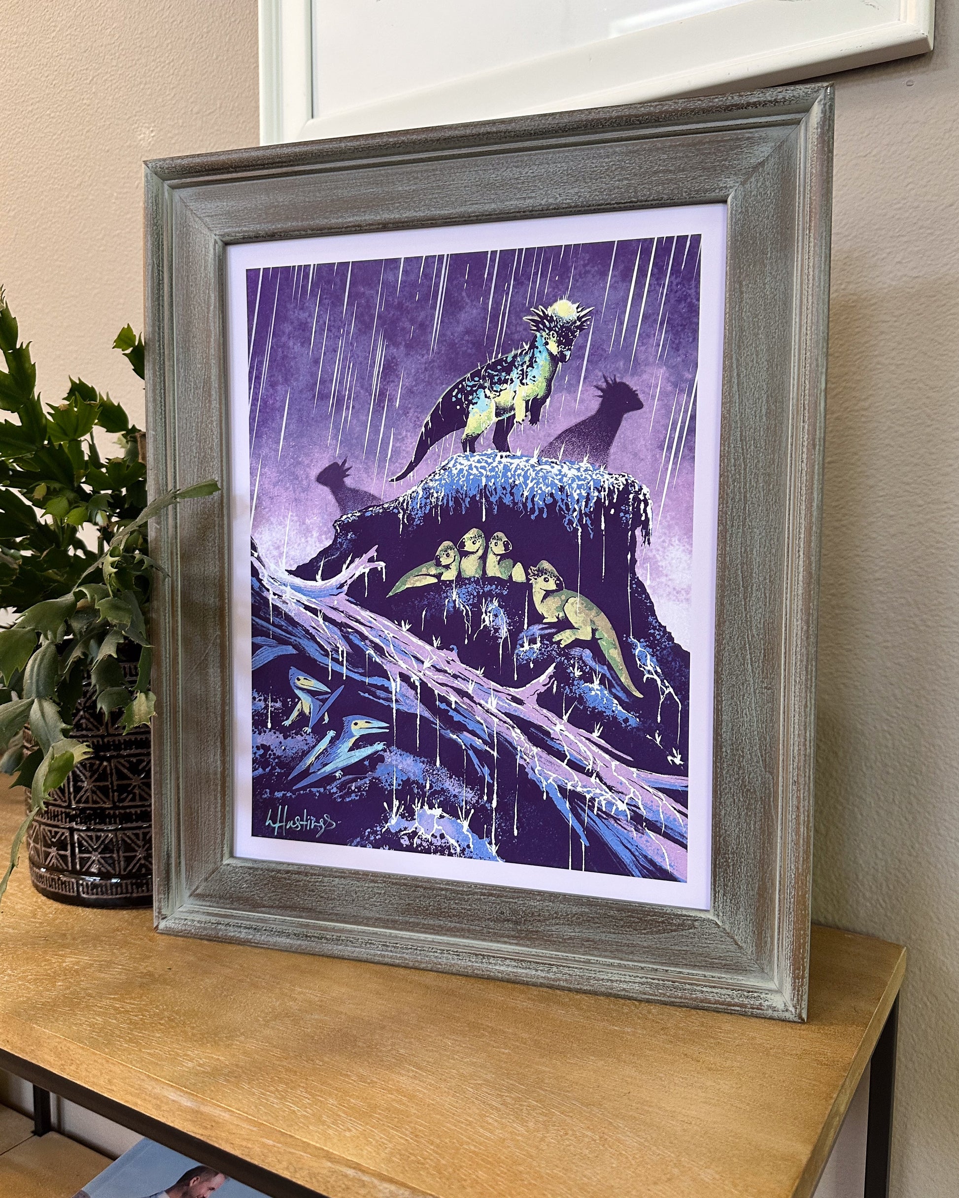 A framed art print featuring stylized dinosaurs in a rainy purple scene, displayed on a wooden table next to a potted plant.