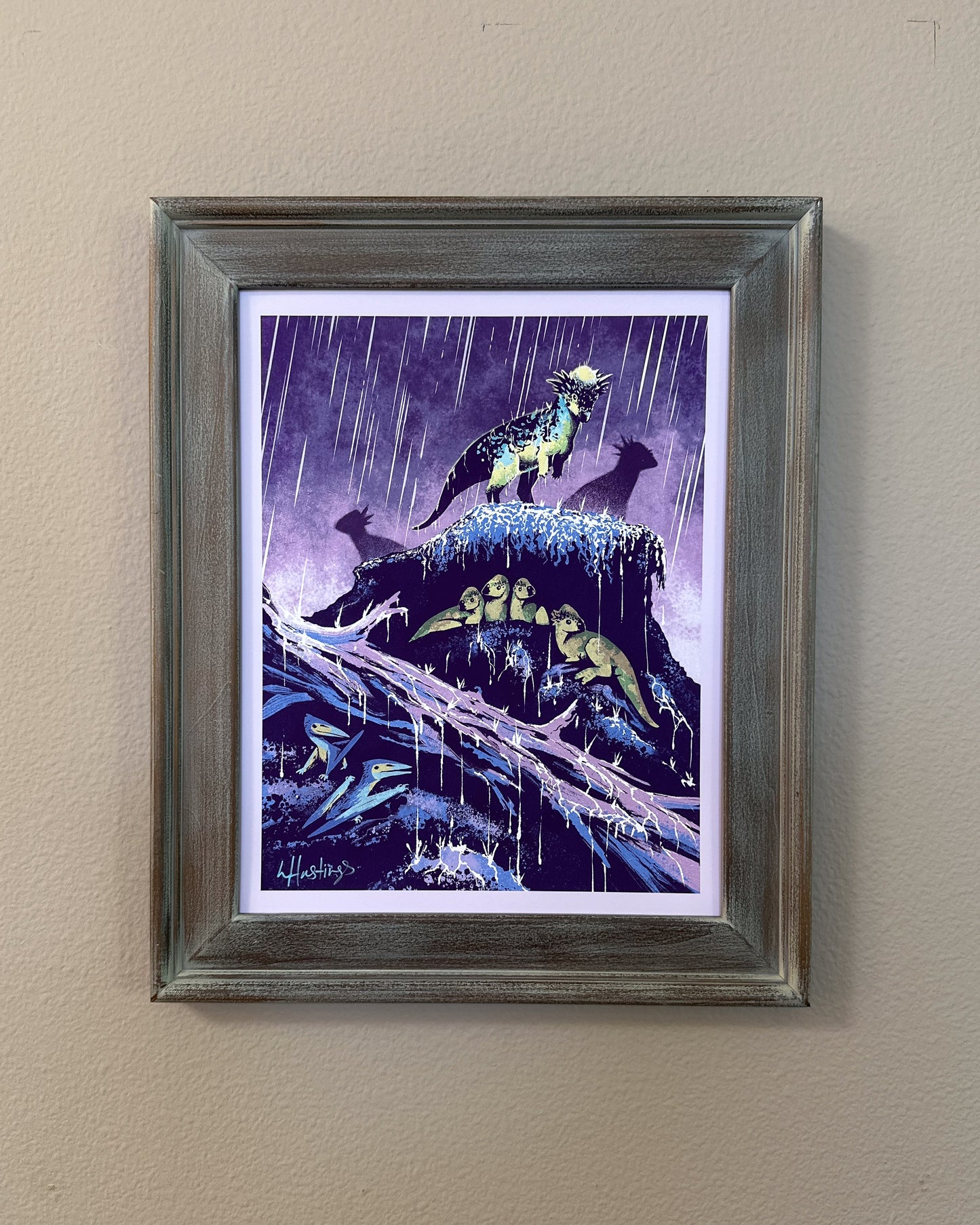 A framed art print featuring a dramatic illustration of dinosaurs in a rainy purple landscape, mounted on a beige wall.