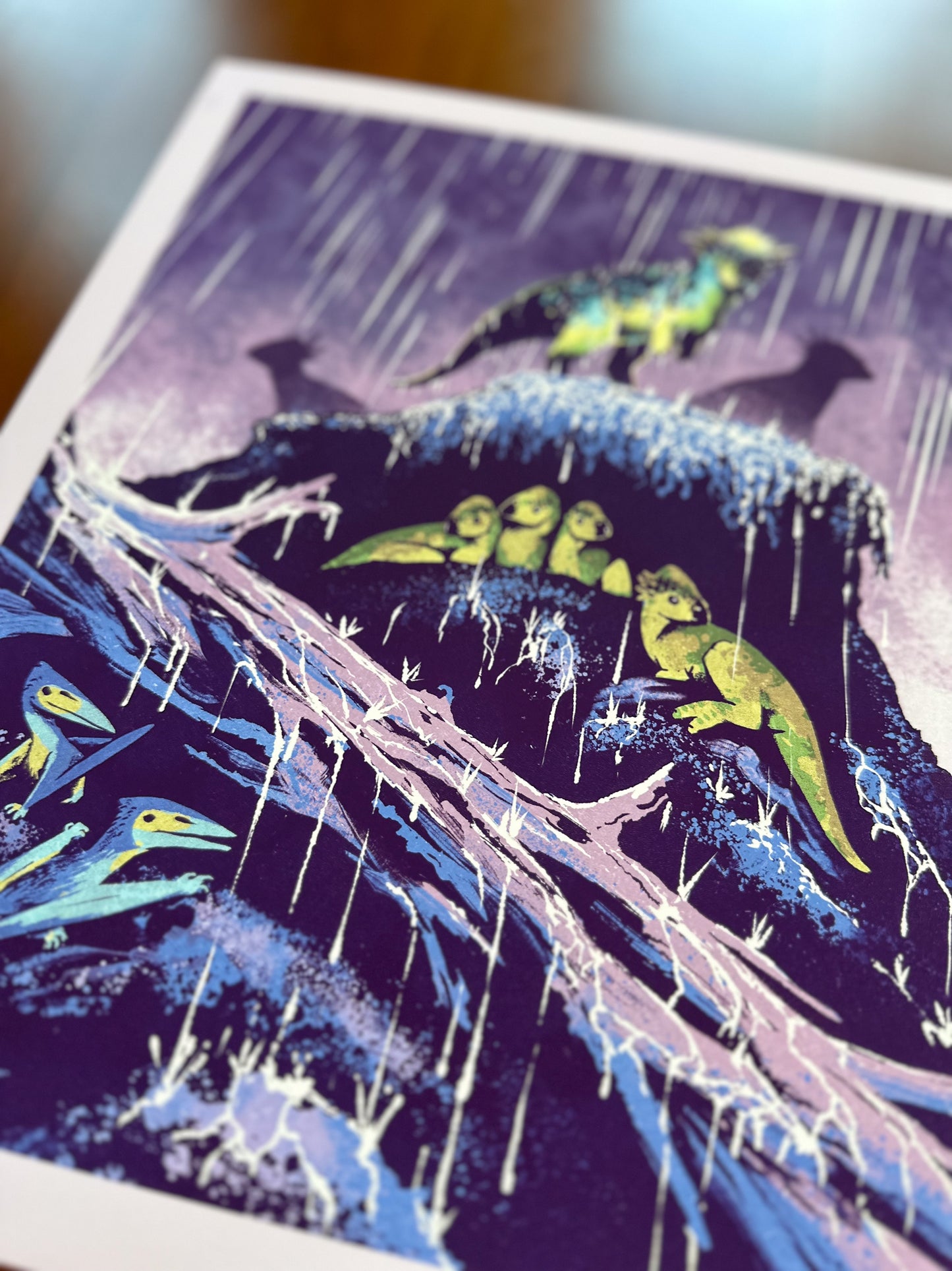 A close-up of an art print featuring a stylized illustration of small dinosaurs on a rainy cliffside under a purple rainy sky, photographed on a wooden surface.