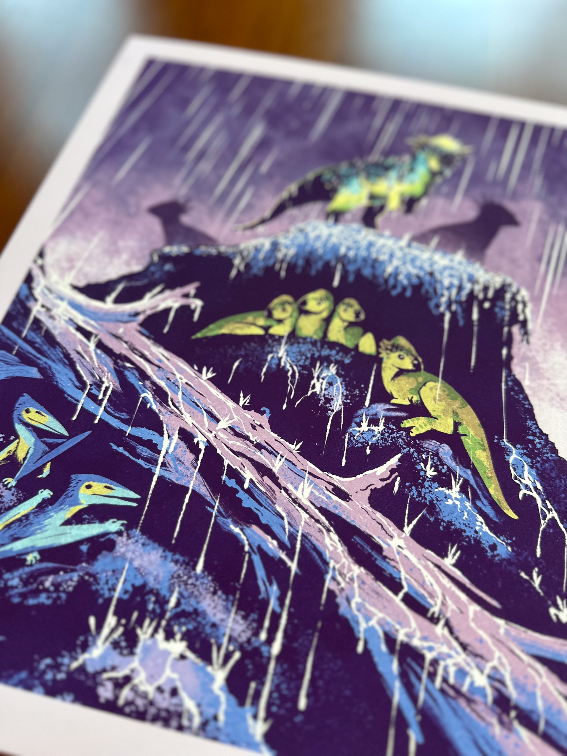 A close-up of an art print featuring a stylized illustration of small dinosaurs on a rainy cliffside under a purple rainy sky, photographed on a wooden surface.